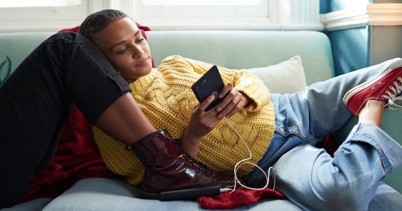 A Black woman relaxes against a friend's legs on the sofa while scrolling through her phone. 