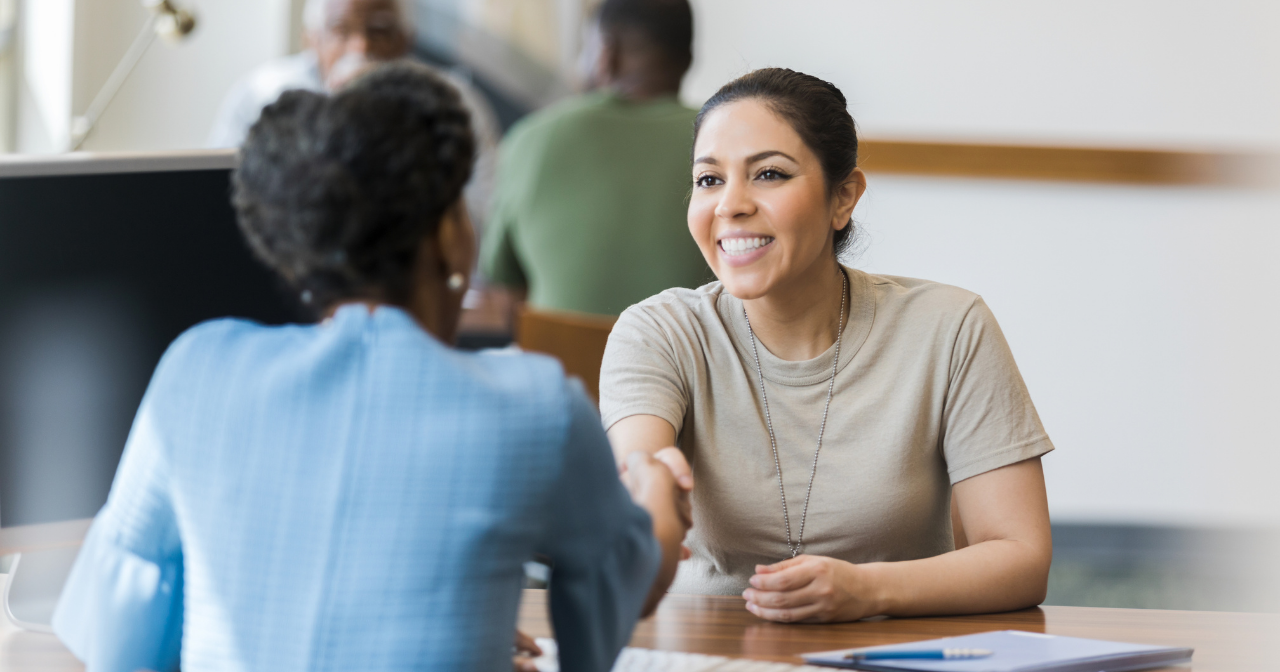 A servicewoman shakes hands with another woman from across a desk. 