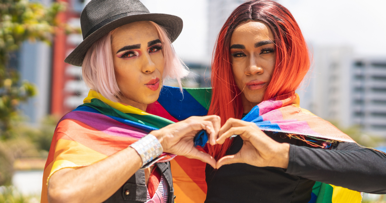 Two queer people wrapped in a rainbow flag make a heart with their hands. 