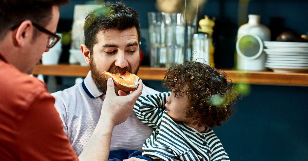 Two dads eat a meal with their young daughter on their lap. 