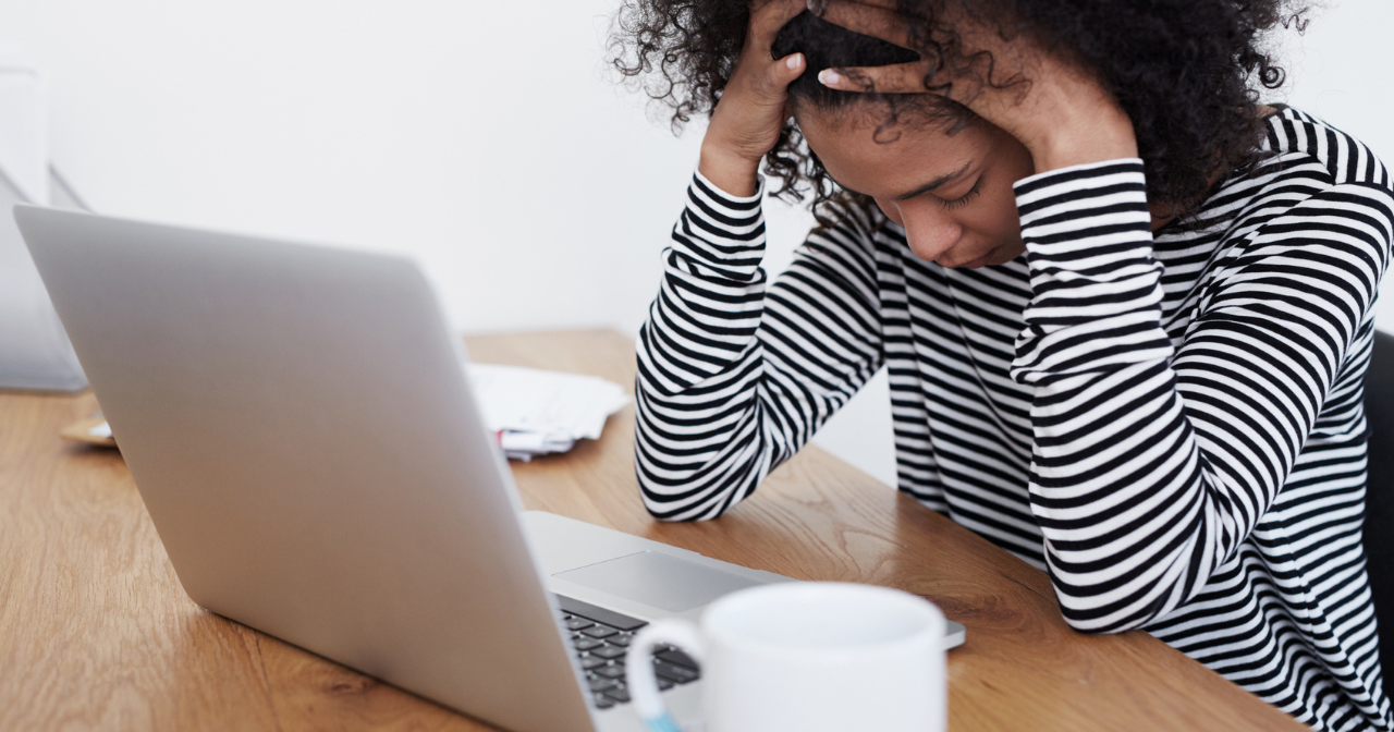 A woman sits at a computer, stressed, with her hands on her head. 