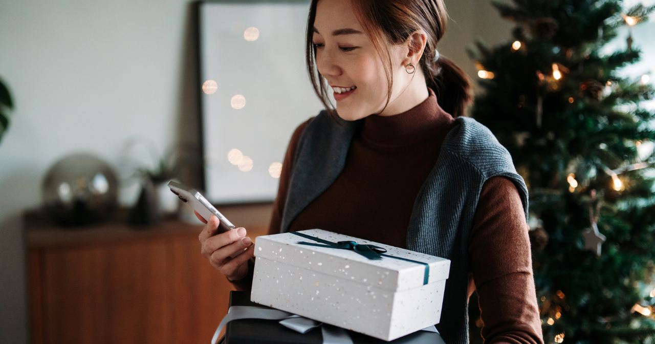 A woman holds a wrapped package in each hand with a Christmas tree in the background.