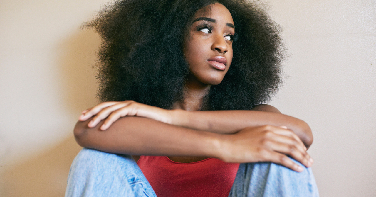 A photo of a Black woman sitting on the floor with her arms crossed, looking concerned. 