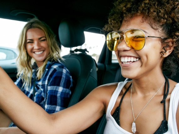 Portrait of two women with long blond and brown curly hair sitting in car, wearing sunglasses, smiling.