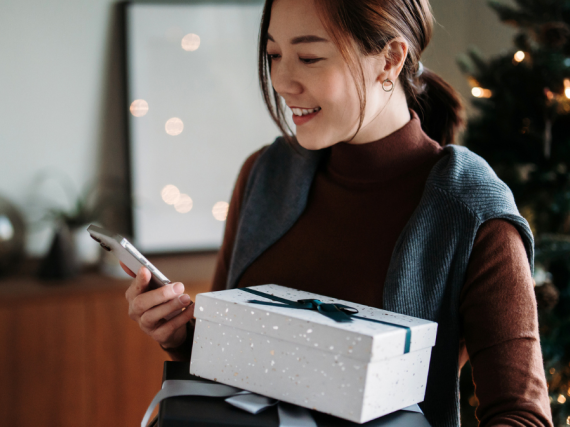 A woman holds a wrapped package in each hand with a Christmas tree in the background.
