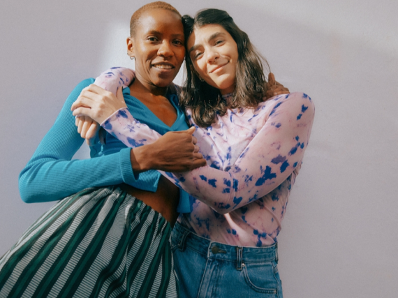 Two women stand against a blank wall and side hug each other. 