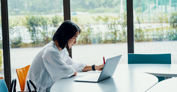 A woman works at her computer while taking notes by hand. 