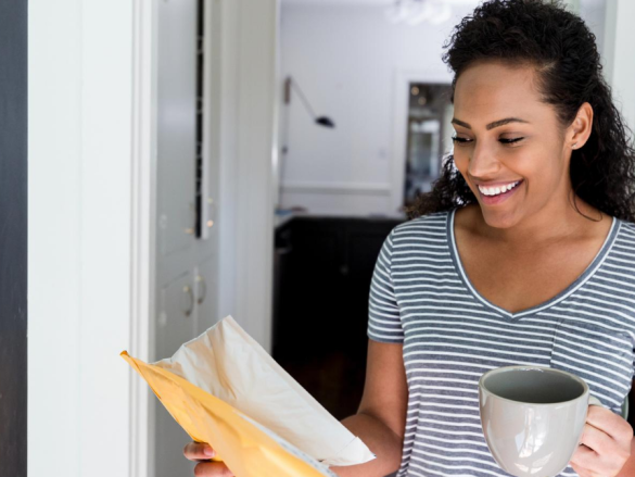 A woman holds a cup of coffee and with a smile picks up a package from her front door. 